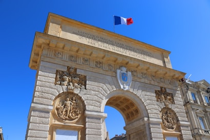 An ornate stone archway displays intricate carvings and sculptures, topped with the French flag against a clear blue sky. Latin inscriptions are engraved on the surface. The structure appears to be part of a historic or monumental building complex.