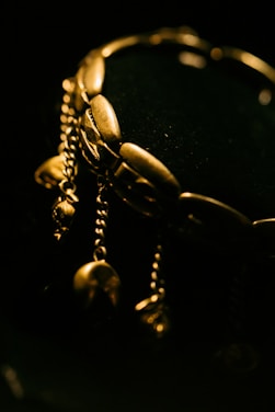 A close-up of an ornate gold bracelet with several dangling chain elements. The bracelets have a lustrous shine against a dark background, which highlights its intricate design.