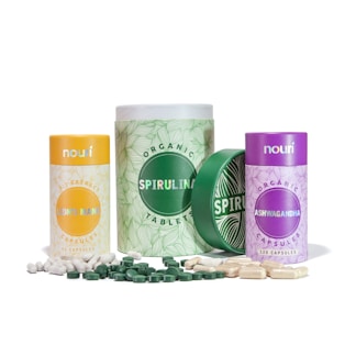 Collection of three cylindrical containers with labels for different dietary supplements. The containers are designed with various colors: yellow for Lion's Mane, green for Spirulina, and purple for Ashwagandha. White and green capsules are scattered around the containers.