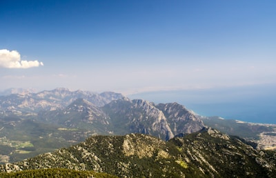 A vast mountain range stretches across the horizon with jagged peaks and ridges covered in greenery. In the background, there is a clear blue sky with a few scattered clouds, and the sea is visible in the distance.
