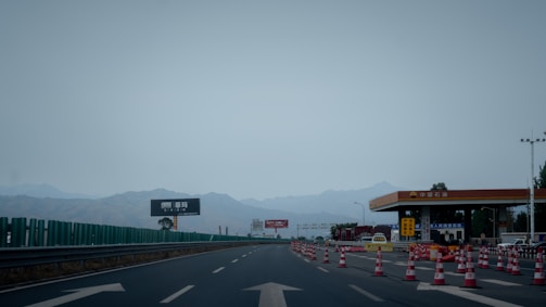 A highway scene with traffic cones lined up to guide vehicles toward a toll station. The toll booth is on the right-hand side with various signs and vehicles present. Mountains form the backdrop, and the sky is overcast.