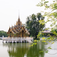An ornate Thai pavilion stands elegantly on a serene lake, its intricate architecture reflecting in the water. Surrounded by lush greenery and tall trees, the scene is tranquil and picturesque. The pavilion’s detailed roof and vibrant colors highlight traditional artistry.