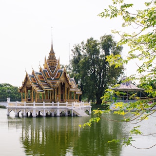 An ornate Thai pavilion stands elegantly on a serene lake, its intricate architecture reflecting in the water. Surrounded by lush greenery and tall trees, the scene is tranquil and picturesque. The pavilion’s detailed roof and vibrant colors highlight traditional artistry.