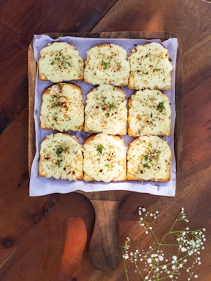 Nine slices of toasted bread topped with melted cheese and sprinkled with herbs and spices are arranged neatly on a wooden serving board. The board is placed on a wooden table, and a few delicate white flowers are visible in the bottom corner.