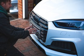 A person crouches in front of a white luxury car, examining or adjusting the license plate on the front bumper. The scene is set on a brick driveway next to a small workshop or garage. The car's grille, LED headlight, and the logo are prominently visible.