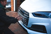 A person crouches in front of a white luxury car, examining or adjusting the license plate on the front bumper. The scene is set on a brick driveway next to a small workshop or garage. The car's grille, LED headlight, and the logo are prominently visible.