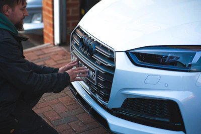 A person crouches in front of a white luxury car, examining or adjusting the license plate on the front bumper. The scene is set on a brick driveway next to a small workshop or garage. The car's grille, LED headlight, and the logo are prominently visible.