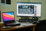 A workspace setup featuring a laptop and a desktop monitor. The laptop displays a colorful abstract background, and the monitor shows a YouTube video with subtitles. A remote, a mug, and some cables are on the wooden desk, with a keyboard and green speakers placed next to the monitor. A window is partially visible in the background.