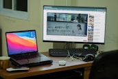 A workspace setup featuring a laptop and a desktop monitor. The laptop displays a colorful abstract background, and the monitor shows a YouTube video with subtitles. A remote, a mug, and some cables are on the wooden desk, with a keyboard and green speakers placed next to the monitor. A window is partially visible in the background.