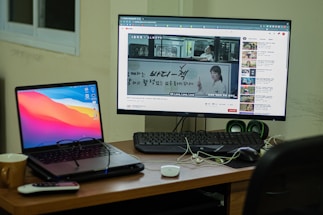 A workspace setup featuring a laptop and a desktop monitor. The laptop displays a colorful abstract background, and the monitor shows a YouTube video with subtitles. A remote, a mug, and some cables are on the wooden desk, with a keyboard and green speakers placed next to the monitor. A window is partially visible in the background.