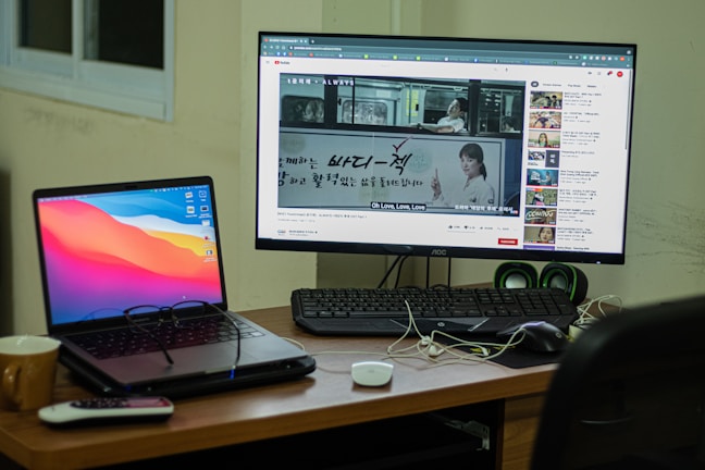 A workspace setup featuring a laptop and a desktop monitor. The laptop displays a colorful abstract background, and the monitor shows a YouTube video with subtitles. A remote, a mug, and some cables are on the wooden desk, with a keyboard and green speakers placed next to the monitor. A window is partially visible in the background.