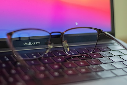 A pair of eyeglasses rests on the keyboard of an open MacBook Pro laptop. The laptop screen displays a gradient of vibrant colors, mainly pinks and purples. The eyeglasses are in focus, while the background is softly blurred, emphasizing the glasses' metal frame.
