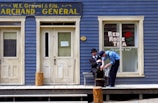 Two people are outside an old-fashioned store with blue wooden siding and cream-colored doors and windows. One person is sitting while the other shines his shoe. The store has a sign saying 'W.E. Gravel & fils. Archand Général' and a window advertisement for 'Red Rose Tea' featuring a red rose graphic.