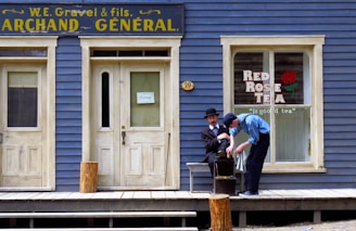 Two people are outside an old-fashioned store with blue wooden siding and cream-colored doors and windows. One person is sitting while the other shines his shoe. The store has a sign saying 'W.E. Gravel & fils. Archand Général' and a window advertisement for 'Red Rose Tea' featuring a red rose graphic.