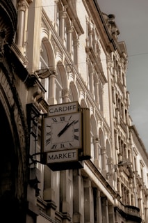 A vintage clock with Roman numerals is mounted on the side of a building, displaying the name 'Cardiff Market.' The architecture features ornate stonework and arched windows, suggesting a historic or classical design. The lighting casts soft shadows, highlighting the textures and details of the facade.