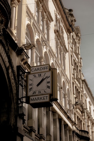 A vintage clock with Roman numerals is mounted on the side of a building, displaying the name 'Cardiff Market.' The architecture features ornate stonework and arched windows, suggesting a historic or classical design. The lighting casts soft shadows, highlighting the textures and details of the facade.