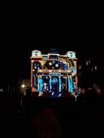 A building is illuminated with colorful, intricate light projections, creating a visual display that resembles a clockwork or machinery theme. The architectural features of the building are accentuated by the light, highlighting columns and intricate designs.