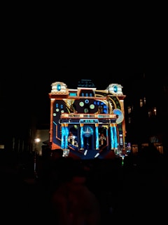 A building is illuminated with colorful, intricate light projections, creating a visual display that resembles a clockwork or machinery theme. The architectural features of the building are accentuated by the light, highlighting columns and intricate designs.