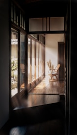 A serene hallway with wooden floors and large windows on one side letting in natural light. Two wicker chairs are positioned at the end of the hallway, and sunlight casts gentle shadows on the floor. The surroundings outside the windows include greenery and rocks.