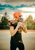 A football player is standing on an outdoor field, wearing an orange helmet and black uniform with orange details. The player is holding a football with both hands, ready to throw. The sky is partly cloudy.