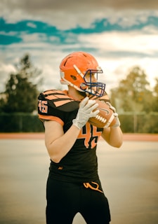 A football player is standing on an outdoor field, wearing an orange helmet and black uniform with orange details. The player is holding a football with both hands, ready to throw. The sky is partly cloudy.