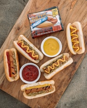 A wooden surface displays several grilled hot dogs in buns, some topped with mustard or ketchup, alongside small bowls of more ketchup and mustard. There's also a package of Ball Park Franks prominently placed on the board.