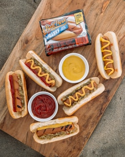 A wooden surface displays several grilled hot dogs in buns, some topped with mustard or ketchup, alongside small bowls of more ketchup and mustard. There's also a package of Ball Park Franks prominently placed on the board.