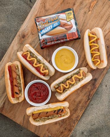 A wooden surface displays several grilled hot dogs in buns, some topped with mustard or ketchup, alongside small bowls of more ketchup and mustard. There's also a package of Ball Park Franks prominently placed on the board.