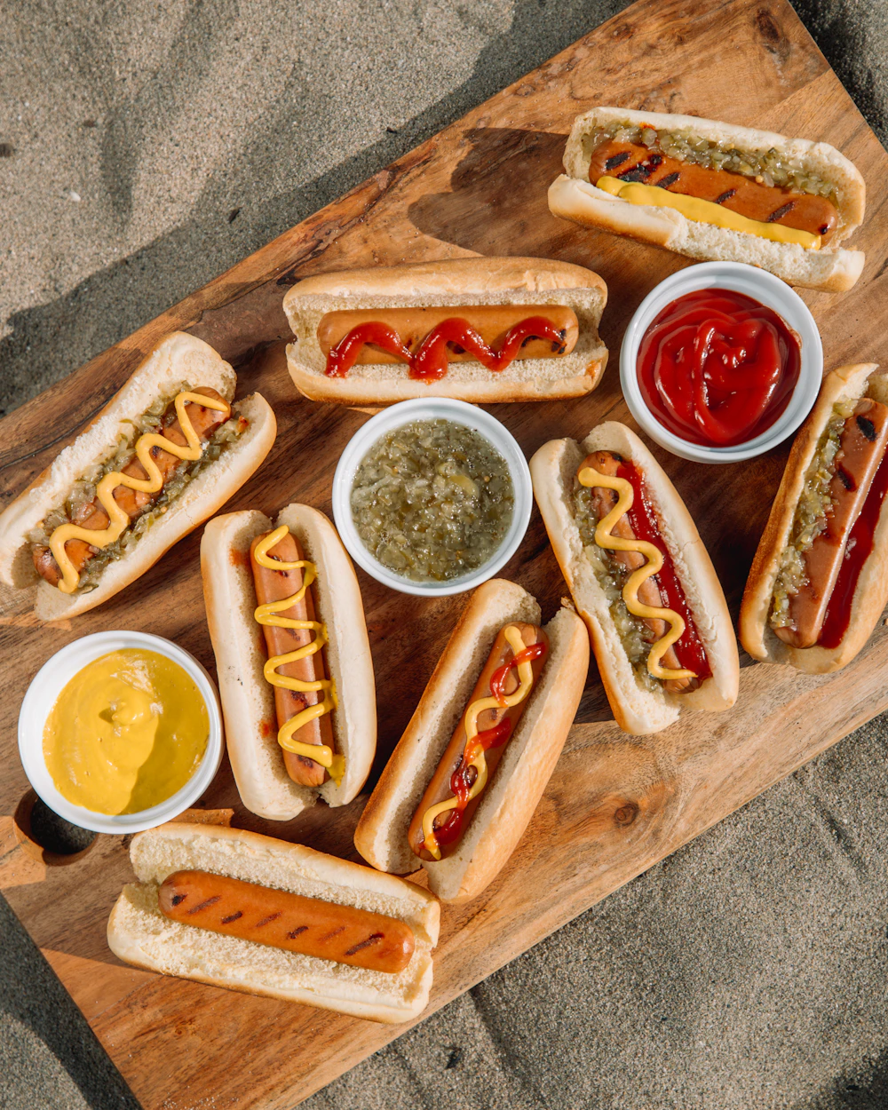 A wooden board lined with hot dogs and small bowls of mustard, ketchup and relish