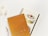 Two spiral-bound notebooks are placed on a light, neutral surface. The first notebook has a golden cover with the text 'TOGETHER is our FAVORITE Place to be' written in a decorative font. The second notebook features an illustration of a cake topped with a cherry and some decorative icing, with text background in a light beige color. To the left of the notebooks, there is a clear pen and two fluffy, white decorative plants.
