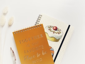 Two spiral-bound notebooks are placed on a light, neutral surface. The first notebook has a golden cover with the text 'TOGETHER is our FAVORITE Place to be' written in a decorative font. The second notebook features an illustration of a cake topped with a cherry and some decorative icing, with text background in a light beige color. To the left of the notebooks, there is a clear pen and two fluffy, white decorative plants.