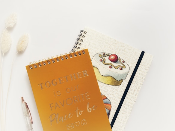 Two spiral-bound notebooks are placed on a light, neutral surface. The first notebook has a golden cover with the text 'TOGETHER is our FAVORITE Place to be' written in a decorative font. The second notebook features an illustration of a cake topped with a cherry and some decorative icing, with text background in a light beige color. To the left of the notebooks, there is a clear pen and two fluffy, white decorative plants.