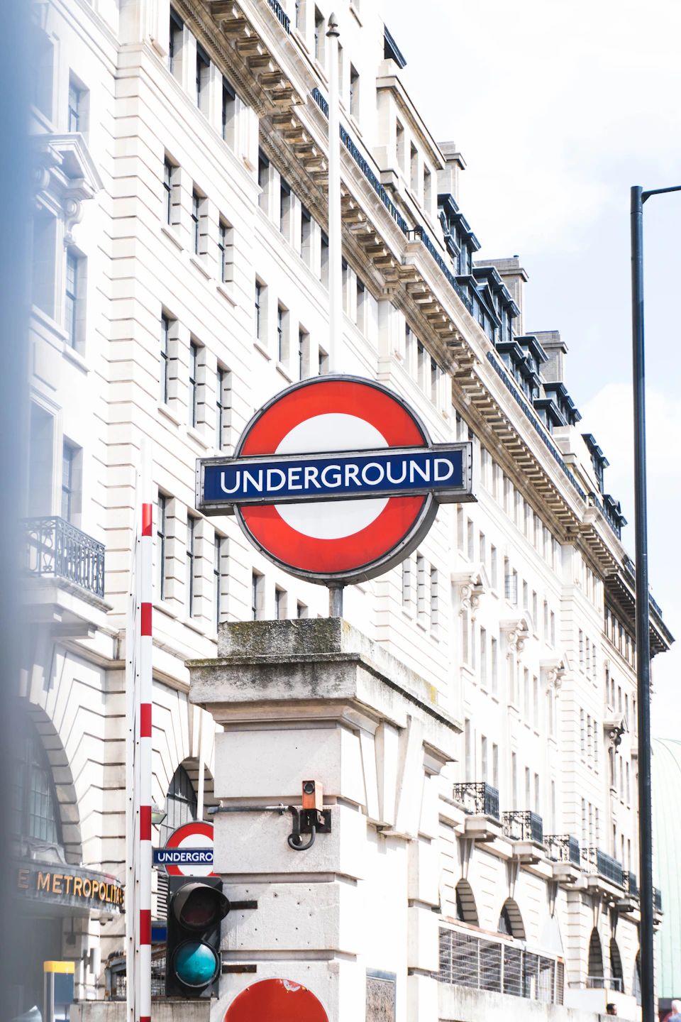 The image shows a street scene with a prominent Underground sign mounted on a white pole, displaying the red and blue logo for the underground transportation system. The pole is located at an intersection, indicating that it serves as a traffic control device to guide vehicles through the area. In front of the pole, there are two traffic lights - one green and one red. On either side of the street, there are buildings with white facades adorned with black roofs, suggesting a cityscape setting.

A few people can be seen walking on the sidewalk in the background, adding life to the scene. The sky above is blue with some clouds scattered across it, indicating that the weather is likely pleasant and sunny.