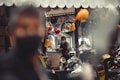 A shop with a variety of hard hats hanging from the ceiling. In the background, two people are seated, both wearing face masks and one is working at a computer. Several scooters are parked in the foreground, with a blurred figure occupying much of the left side of the frame. The interior details suggest an industrial or construction-related environment.