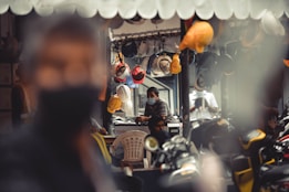 A shop with a variety of hard hats hanging from the ceiling. In the background, two people are seated, both wearing face masks and one is working at a computer. Several scooters are parked in the foreground, with a blurred figure occupying much of the left side of the frame. The interior details suggest an industrial or construction-related environment.