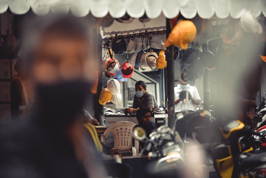 A shop with a variety of hard hats hanging from the ceiling. In the background, two people are seated, both wearing face masks and one is working at a computer. Several scooters are parked in the foreground, with a blurred figure occupying much of the left side of the frame. The interior details suggest an industrial or construction-related environment.