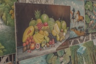 A collection of paintings is displayed, with the central artwork being a still life of a variety of fruits including bananas, pineapples, watermelon, and others arranged on a table. Surrounding it are paintings of different subjects such as landscapes with waterfalls, horse riders, and abstract art featuring fish and lotus leaves.