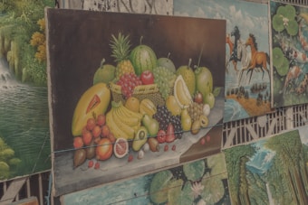 A collection of paintings is displayed, with the central artwork being a still life of a variety of fruits including bananas, pineapples, watermelon, and others arranged on a table. Surrounding it are paintings of different subjects such as landscapes with waterfalls, horse riders, and abstract art featuring fish and lotus leaves.