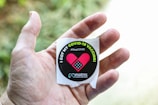 A hand holding a circular sticker with a message indicating receipt of the COVID-19 vaccine. The sticker features a pink heart with a bandage and the text 'I Got My COVID-19 Vaccine' along with '#StopCOVID' printed on it. The background is blurred with green foliage visible.
