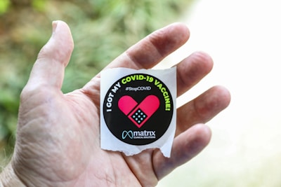A hand holding a circular sticker with a message indicating receipt of the COVID-19 vaccine. The sticker features a pink heart with a bandage and the text 'I Got My COVID-19 Vaccine' along with '#StopCOVID' printed on it. The background is blurred with green foliage visible.
