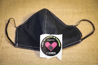 A black face mask is placed on a textured beige surface. In front of the mask, there is a round sticker with the text 'I got my COVID-19 vaccine' and '#StopCOVID'. The sticker features an image of a pink bandage and the name 'matrix' below it.