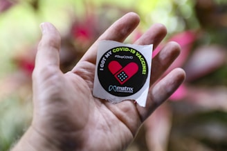 A hand holds a circular sticker that reads 'I GOT MY COVID-19 VACCINE' with the hashtag #StopCOVID. The sticker features a red heart symbol with a small band-aid graphic. The background is blurred with hints of green and pink, suggesting an outdoor setting.