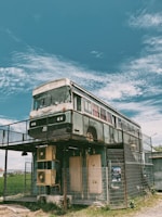 A double-decker bus has been creatively repurposed as a two-story structure. It stands on stilts and is enclosed by a metal fence. The sky above is clear with light, wispy clouds, and there's a grassy area surrounding the structure.