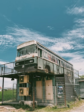 A double-decker bus has been creatively repurposed as a two-story structure. It stands on stilts and is enclosed by a metal fence. The sky above is clear with light, wispy clouds, and there's a grassy area surrounding the structure.