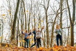 Children joyfully play in a forest during autumn, surrounded by tall trees with sparse leaves and a ground covered in fallen golden leaves. The atmosphere is lively as the children engage with their environment, expressing happiness and energy.