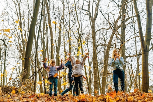 Children joyfully play in a forest during autumn, surrounded by tall trees with sparse leaves and a ground covered in fallen golden leaves. The atmosphere is lively as the children engage with their environment, expressing happiness and energy.