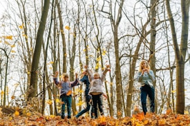 Children joyfully play in a forest during autumn, surrounded by tall trees with sparse leaves and a ground covered in fallen golden leaves. The atmosphere is lively as the children engage with their environment, expressing happiness and energy.