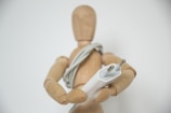 A wooden mannequin holds an electrical plug, with a cord wrapped around its torso. The mannequin is posed against a plain, neutral background.