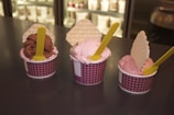 Three cups of ice cream are displayed on a dark countertop. Each cup contains a scoop of ice cream with a waffle biscuit inserted vertically and a yellow spoon resting on the surface. The ice cream flavors appear to include chocolate and strawberry, and the cups are patterned with a red and white design.