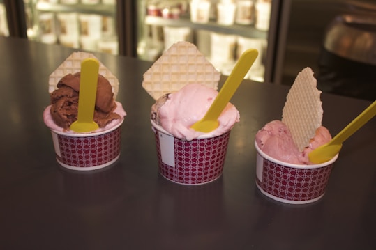 Three cups of ice cream are displayed on a dark countertop. Each cup contains a scoop of ice cream with a waffle biscuit inserted vertically and a yellow spoon resting on the surface. The ice cream flavors appear to include chocolate and strawberry, and the cups are patterned with a red and white design.